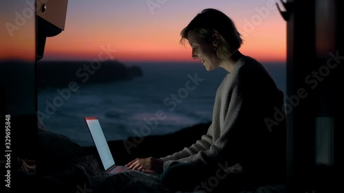 A young entrepreneur woman is working on her computer in a camper van. The door is open to an epic view of the sea in a sunset / sunrise. The lighthouse is in the background. Remote working anywhere.