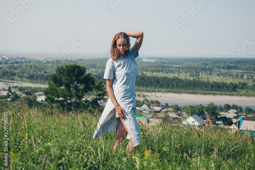 a girl in a loose dress with loose hair walks in a meadow. Village houses, forest and river as background. The concept of summer, warmth, freedom, village life, sunburn