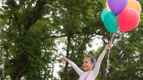 happy little asian girl child playing with colorful toy balloons outdoors. trees and green gardens background. smiling girl with balloons on the street in the summer. freedom and imagination concept.