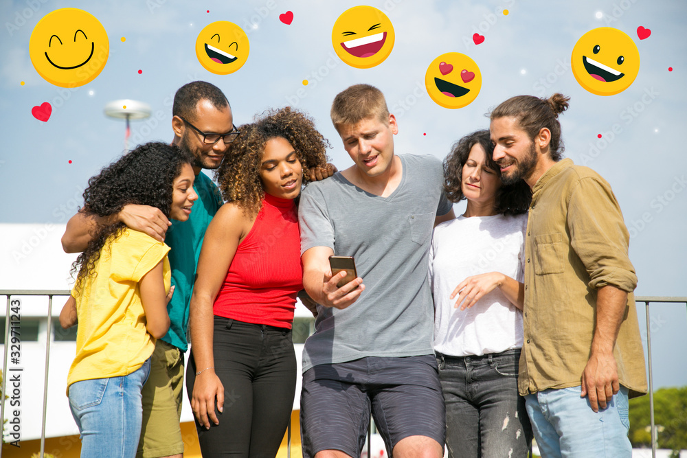 Excited friends watching adorable photos on smartphone with online chat icons. Young Caucasian man holding phone, mix raced group of people looking at screen with cute faces. Using phone concept