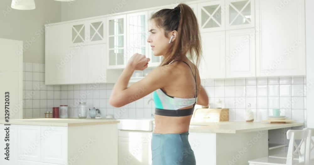 Sporty woman making abs exercises crunches standing in kitchen at home. Girl in sportswear exercising abdominals and listening music in earphones. Sport, fitness, training, workout, wellness concept. Sporty woman making abs exercises crunches standing in kitchen at home. Girl in sportswear exercising abdominals and listening music in earphones. Sport, fitness, training, workout, wellness concept.