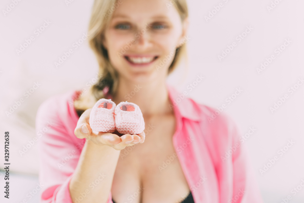 Joyful future mom expecting daughter. Cheerful young woman holding and showing pink baby shoes and smiling. Anticipation concept Joyful future mom expecting daughter. Cheerful young woman holding and showing pink baby shoes and smiling. Anticipation concept