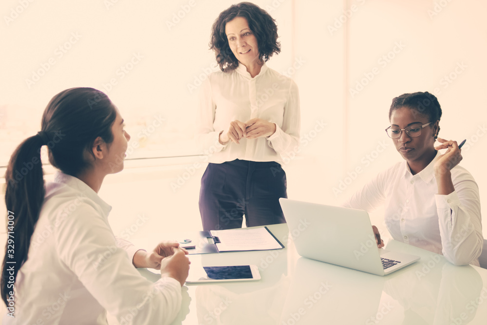 Female business leader explaining project specific to her team. Businesswomen sitting and standing at table, using laptop and talking. Corporate meeting concept Female business leader explaining project specific to her team. Businesswomen sitting and standing at table, using laptop and talking. Corporate meeting concept