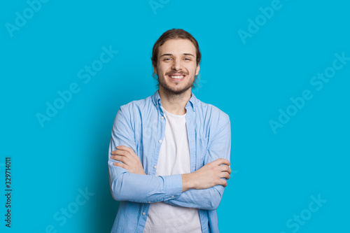 Handsome caucasian man with beard and long hair is posing with crossed hands on a blue background