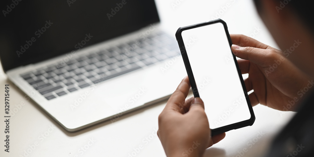 Cropped image of businessman's hands holding a cropped black smartphone with white blank screen putting on modern white table in front computer laptop. Cropped image of businessman's hands holding a cropped black smartphone with white blank screen putting on modern white table in front computer laptop.