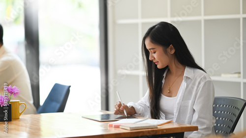 Photo of beautiful woman in white shirt and vest writing on computer tablet with stylus pen while sitting at the modern wooden table with orderly office shelf as background.