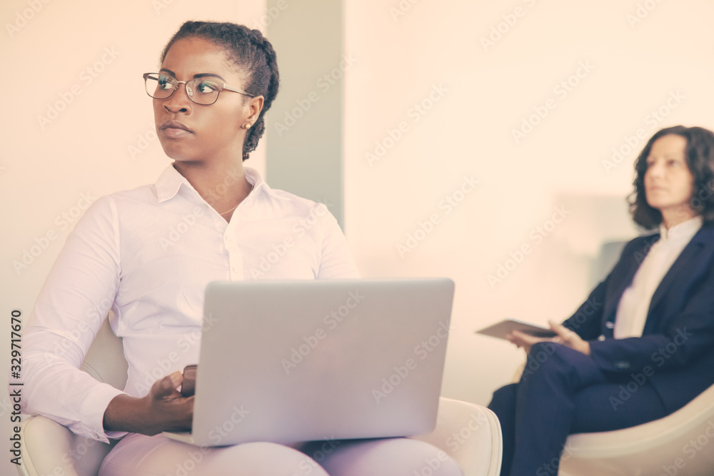 Serious female office worker working in office lounge. Diverse business women sitting in armchairs, using smartphone and laptop in office lobby. Communication concept Serious female office worker working in office lounge. Diverse business women sitting in armchairs, using smartphone and laptop in office lobby. Communication concept