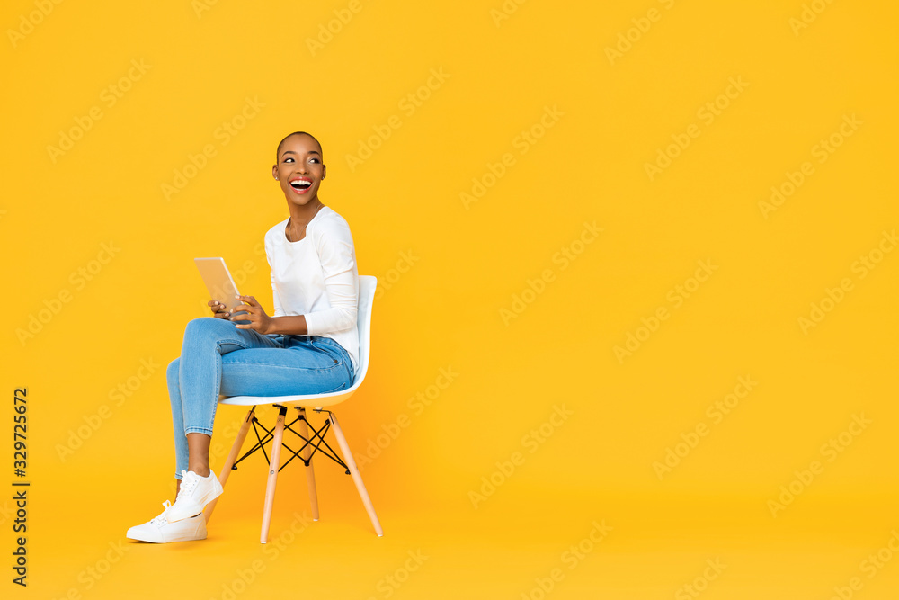 Trendy smiling African American woman sitting on a chair using tablet computer thinking and looking at empty space aside isolated yellow background Trendy smiling African American woman sitting on a chair using tablet computer thinking and looking at empty space aside isolated yellow background