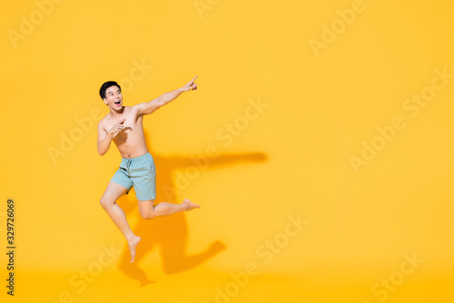 Full length portrait of young happy shirtless Asian man in beach attire jumping in mid-air pointing two fingers at blank space beside in yellow isolated studio background