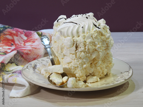 Delicious meringue cake in a white vintage plate with a spoon and napkin on a light wooden table close up. Sweet pastry made of beaten eggs and butter cream.