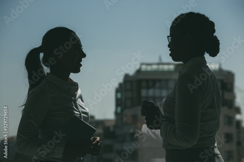 Silhouettes of female colleagues chatting near office window. Businesswomen standing against urban view, talking and smiling. Business colleagues concept