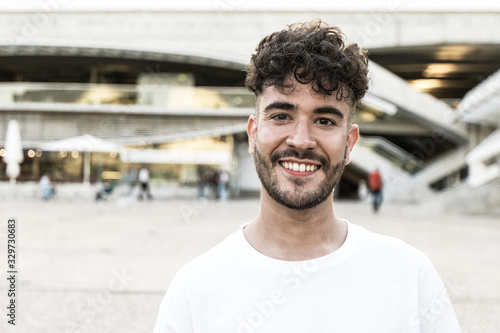 Happy carefree guy posing in city square. Front portrait on young curly haired man in white casual shirt looking and smiling at camera. Male portrait concept