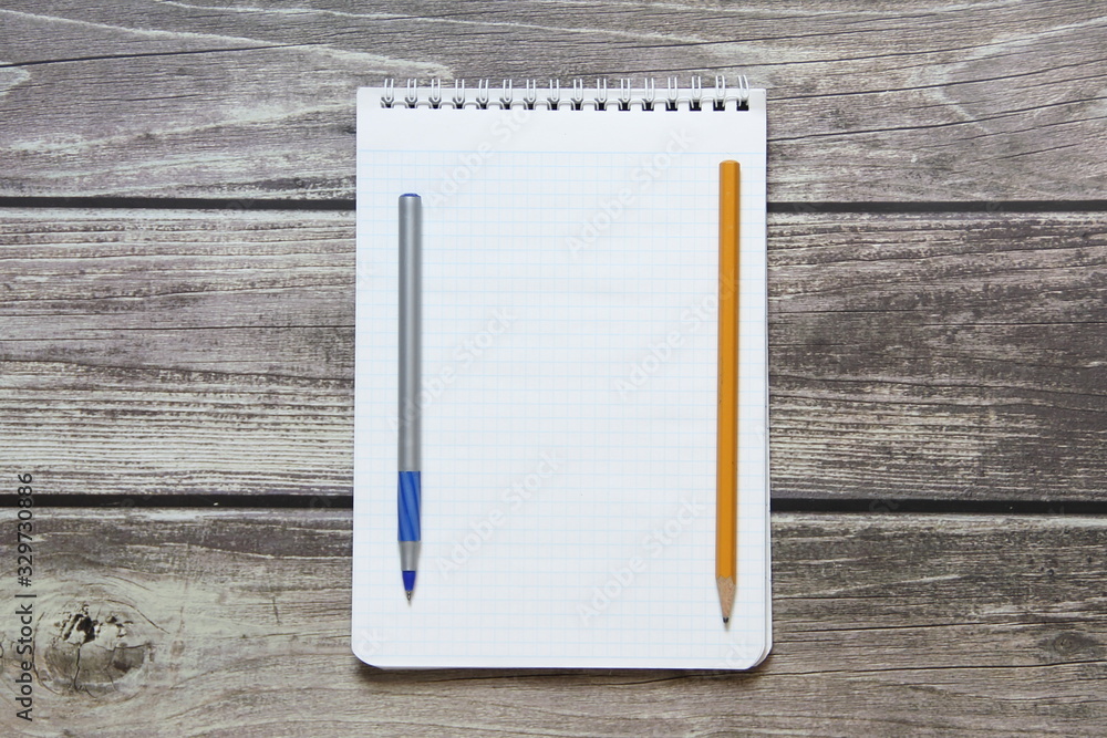 Notepad with a blank white sheet in a checker paper with ball pen and simple graphite pencil lies on the background of wooden boards. Notepad with a blank white sheet in a checker paper with ball pen and simple graphite pencil lies on the background of wooden boards.
