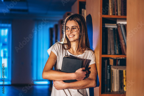 Young girl student with glasses in library stands near bookshelves. Girl smiles and holds a book in her hands. Exam preparation