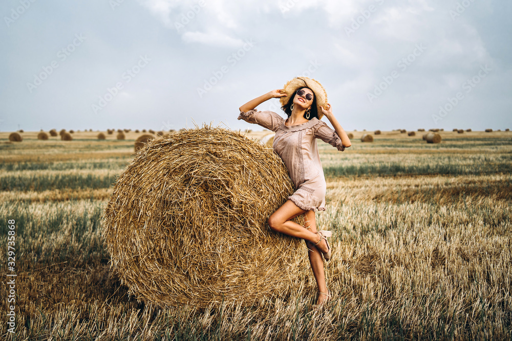 Beautiful young girl with long hair in sunnglasses and straw hat posing on a wheat field near hay bales. Happy brunette in summer dress Beautiful young girl with long hair in sunnglasses and straw hat posing on a wheat field near hay bales. Happy brunette in summer dress