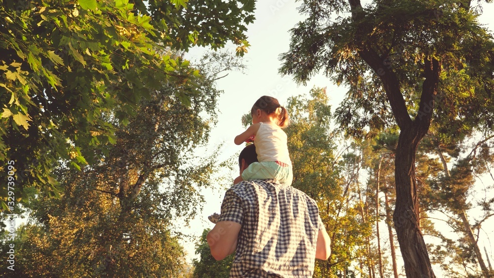 Dad carries on shoulders of his beloved child, in park. Father walks with his daughter on his shoulders under trees. child with parents walks on day off. Happy family is relaxing in park.