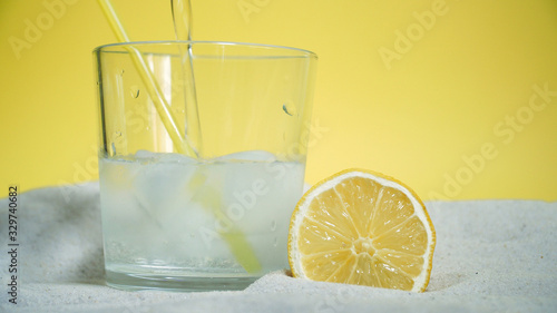 glass with ice and soda water and lemon on the sand on a yellow background