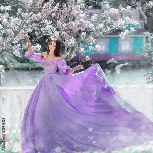 Portrait of amazing cute young woman with dark brown long hair in long purple dress with large flyingskirts stands under blooming white magnolia flowers against background of lake and blue fairy house