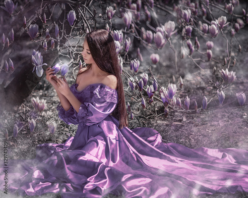 Portrait of amazing cute young woman with dark brown long hair in long purple dress sitting under blooming lilac magnolia tree and holding flower in her hands