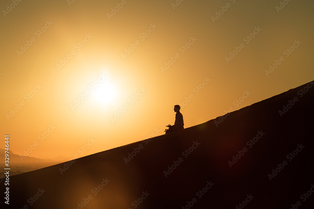 Silhoutte conceptual shot of young man meditating in the desert during sunrise on a sand dune; peaceful meditation in isolated place 