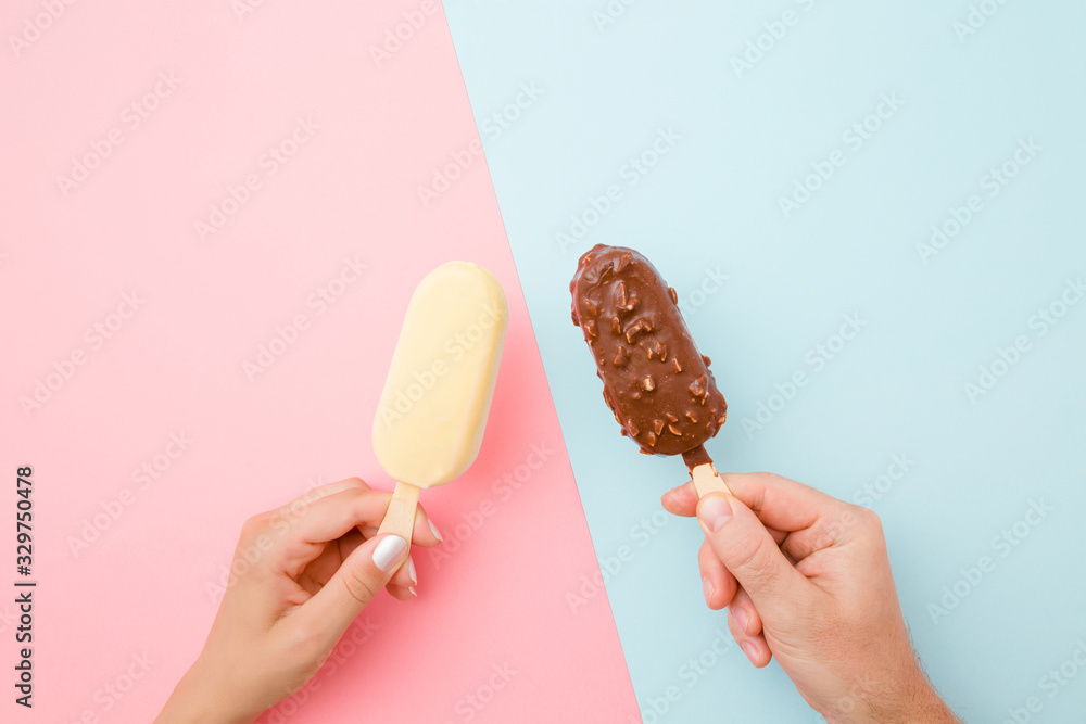 Young couple hands holding together ice creams with dark and white chocolate glaze. Closeup. Point of view shot. Light pink and blue table background. Pastel colors. Top down view. Young couple hands holding together ice creams with dark and white chocolate glaze. Closeup. Point of view shot. Light pink and blue table background. Pastel colors. Top down view.