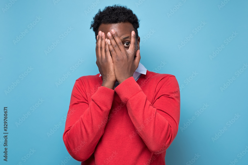 Young african american man in red sweater isolated on blue studio background peeping through fingers, hiding cover face with hands, looking terrified Young african american man in red sweater isolated on blue studio background peeping through fingers, hiding cover face with hands, looking terrified