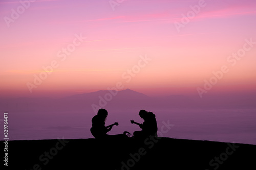 A couple of musicians play the guitar at sunset. Shilhouette of two guys with their guitars next to the see in a pink sunset.