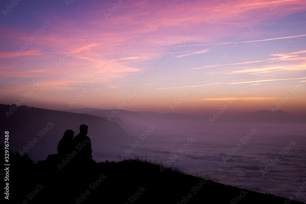 A couple hugs in a romantic sunset at the top of a cliff imaging a future together while looking at the sun