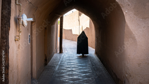 Unidentified muslim woman  dressed in black hijab walks on the narrow street of old city Yazd  Iran.