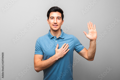 I swear. handsome young man in blue shirt with hand on chest giving oath against grey background. studio shoot