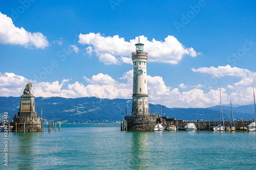 Lindau, Bavaria, Germany. Old lighthouse with clock in bay. Antique bavarian town at Lake Constance Bodensee. Monument with statue of lion at entrance to port, yachts by piers. Summer landscape. © annaartday