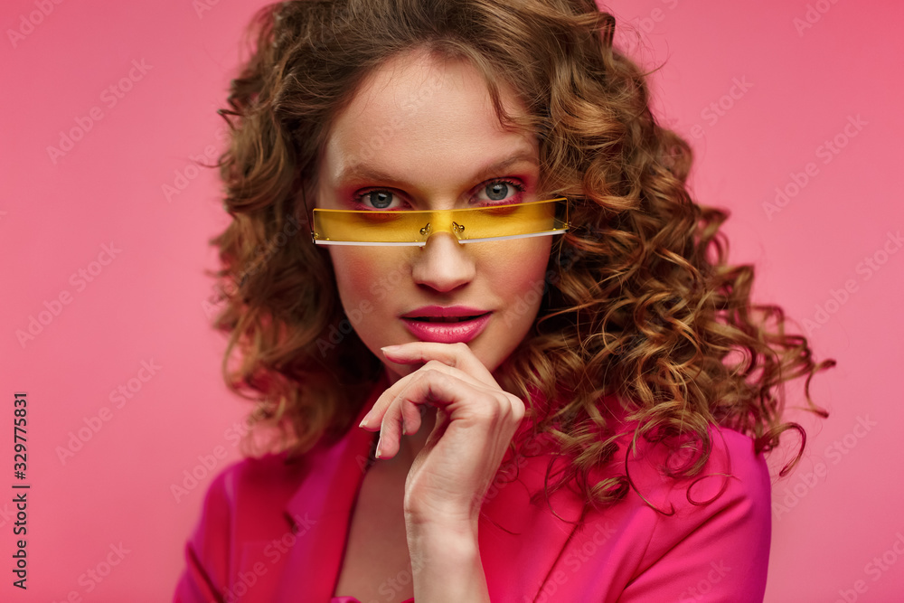 Close up fashion studio portrait of young beautiful women with curly hair.Nice girl with wavy hairstyle,bright make up and stylish eyeglasses isolated over pink background.Beauty and hair care concept