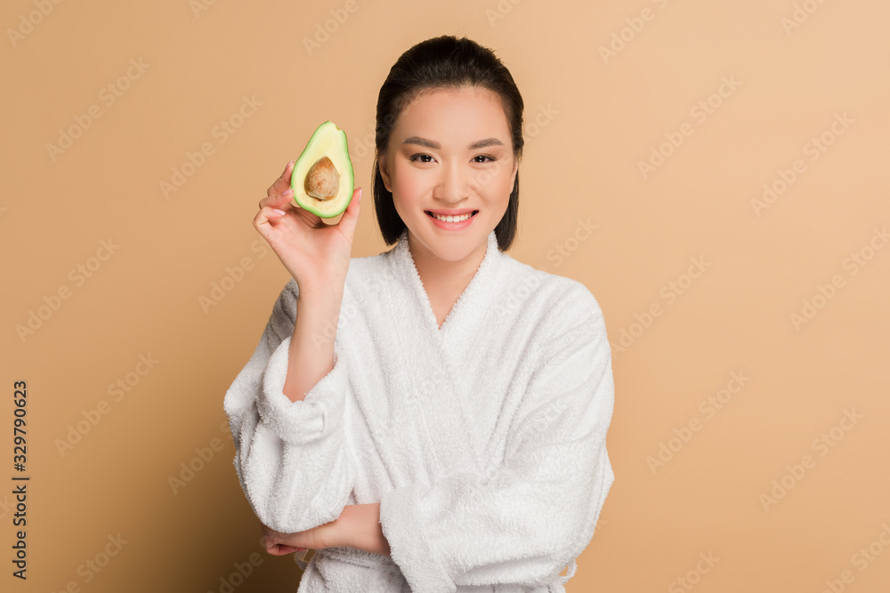 happy beautiful asian woman in bathrobe with avocado half on beige background happy beautiful asian woman in bathrobe with avocado half on beige background