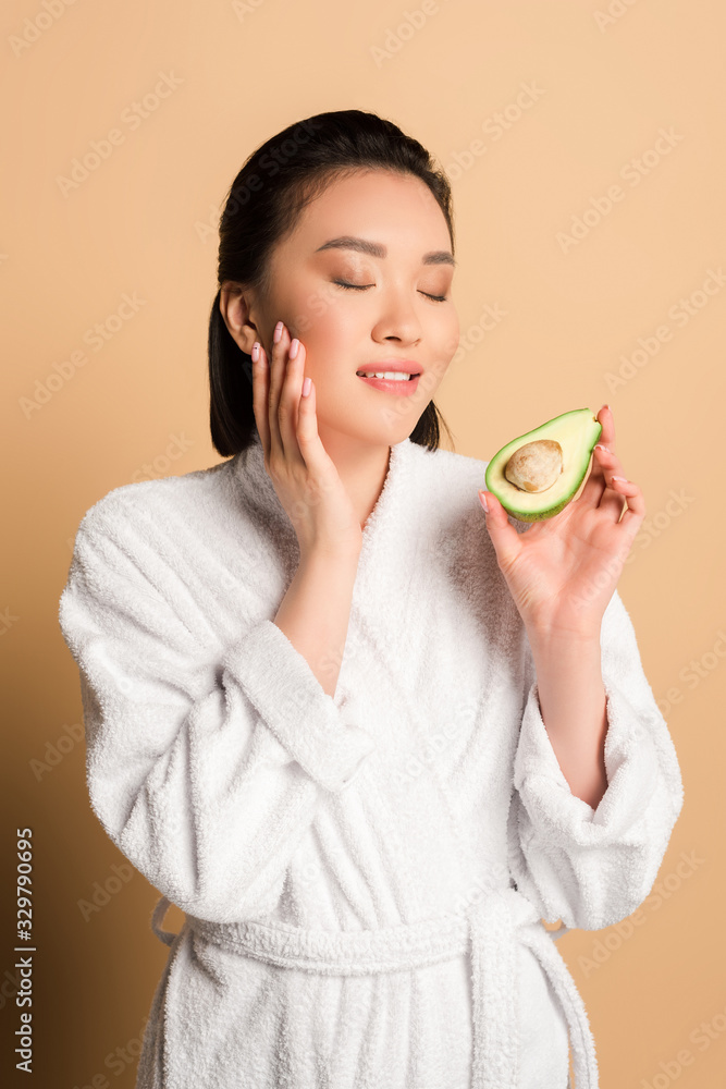 beautiful asian woman in bathrobe with avocado half touching face on beige background beautiful asian woman in bathrobe with avocado half touching face on beige background