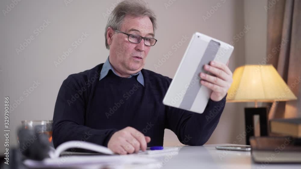 Senior Caucasian man writing at the table when having incoming video call. Mature male retiree in eyeglasses waving and talking at selfie camera. Lifestyle, modern technologies, communication. Senior Caucasian man writing at the table when having incoming video call. Mature male retiree in eyeglasses waving and talking at selfie camera. Lifestyle, modern technologies, communication.