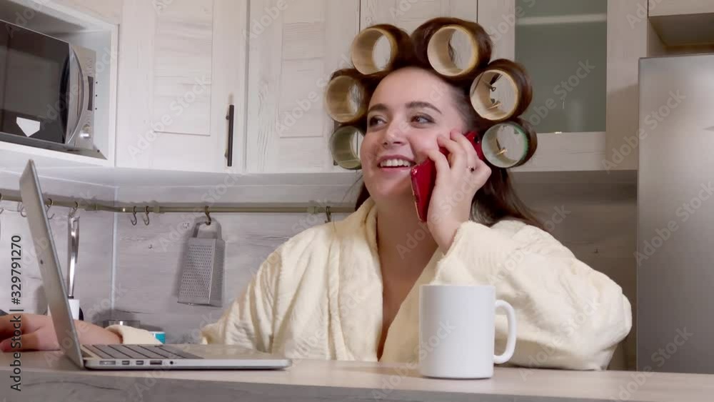 Plus-size girl, dressed in a bathrobe, curlers on her head, talking by phone sitting at kitchen Plus-size girl, dressed in a bathrobe, curlers on her head, talking by phone sitting at kitchen