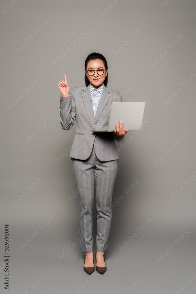 smiling young businesswoman in eyeglasses using laptop and showing idea gesture on grey background smiling young businesswoman in eyeglasses using laptop and showing idea gesture on grey background