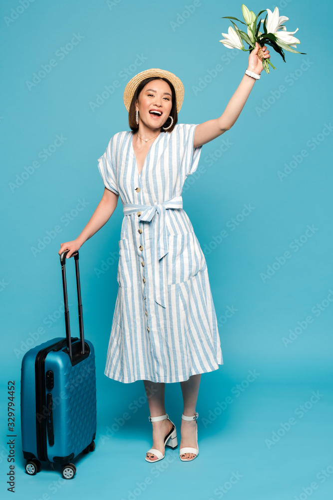 happy brunette asian girl in striped dress and straw hat with travel bag and bouquet of lilies on blue background happy brunette asian girl in striped dress and straw hat with travel bag and bouquet of lilies on blue background