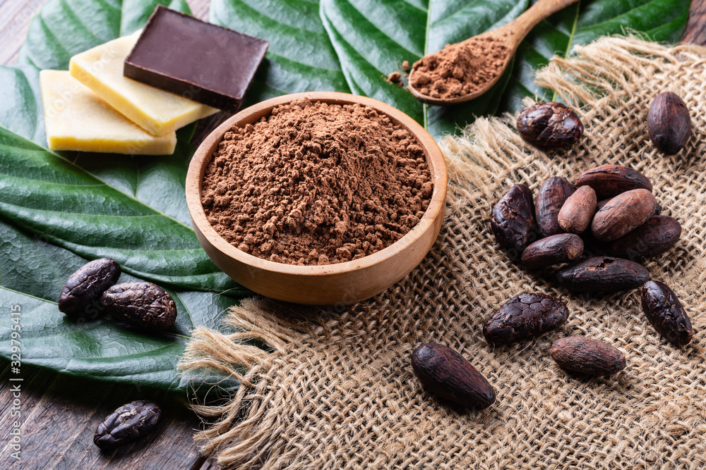 Cocoa powder in wooden bowl, bars of dark chocolate and cocoa butter with whole cacao beans  and fresh leaves on old rustic table.