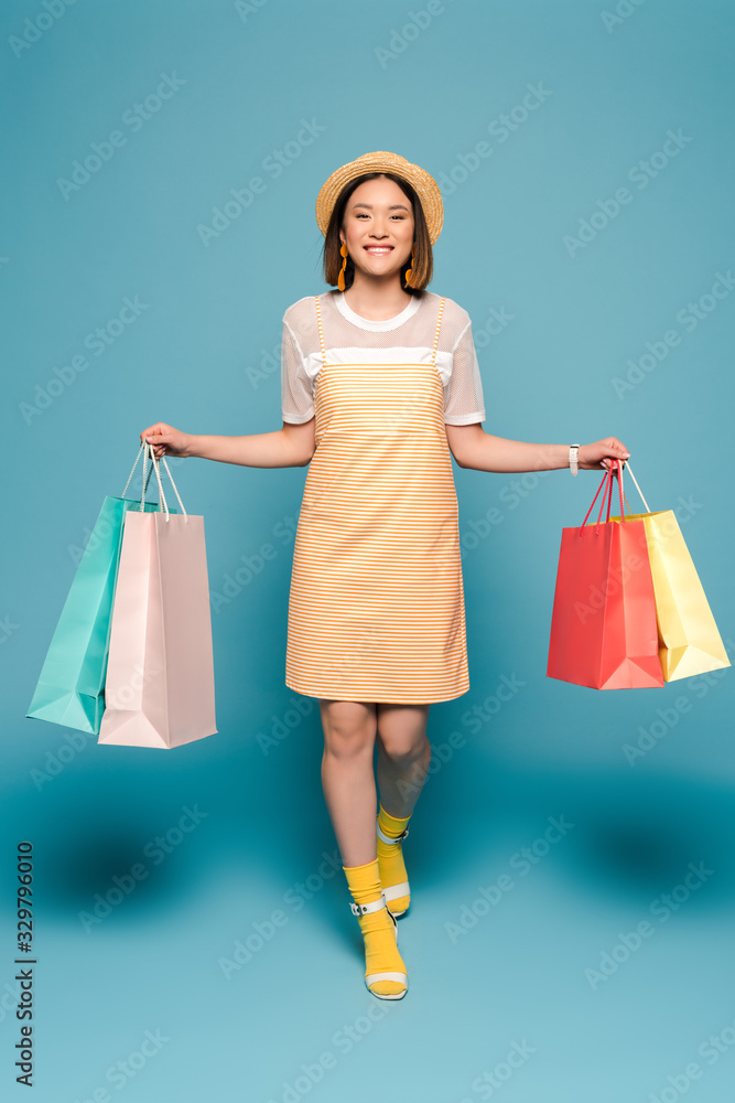 smiling asian girl in striped yellow dress and straw hat with shopping bags on blue background