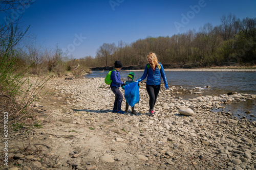 young blond hair woman with her sons in blue gloves cleaning the river bank from plastic and garbage in big blue trash bags on a sunny day in early spring