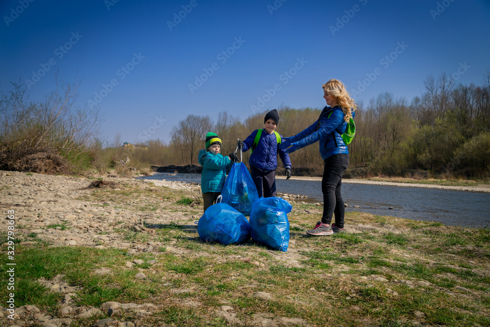 young blond hair woman with her sons in blue gloves cleaning the river bank from plastic and garbage in big blue trash bags on a sunny day in early spring young blond hair woman with her sons in blue gloves cleaning the river bank from plastic and garbage in big blue trash bags on a sunny day in early spring