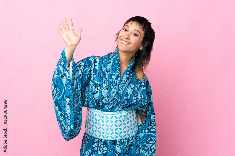 Young woman wearing kimono over isolated blue background saluting with hand with happy expression Young woman wearing kimono over isolated blue background saluting with hand with happy expression