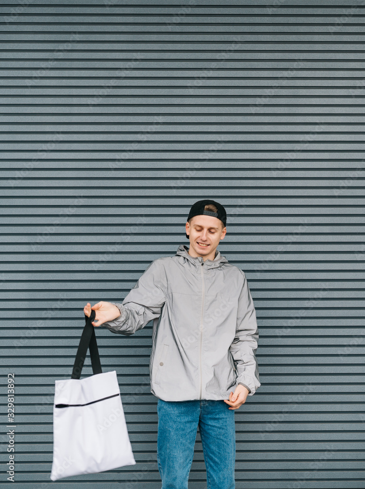 Φωτογραφία Smiling young man in stylish casual clothes stands with a white shopping bag in his hand on a gray background and looks into the camera with a smile on his face