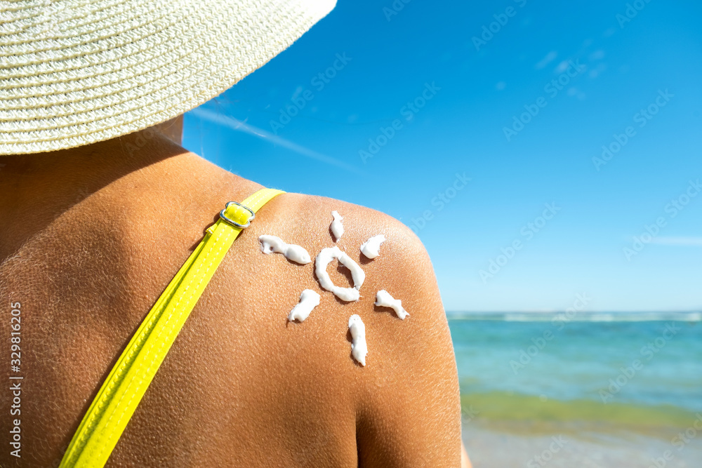Fototapeta premium Back view of young woman tanning at the beach with sunscreen cream in sun shape on her shoulder. UV sunburn protection and sunblock skincare concept