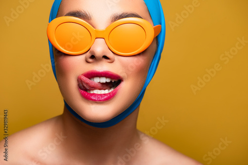 happy girl in scarf and fashionable sunglasses licking lip, isolated on yellow