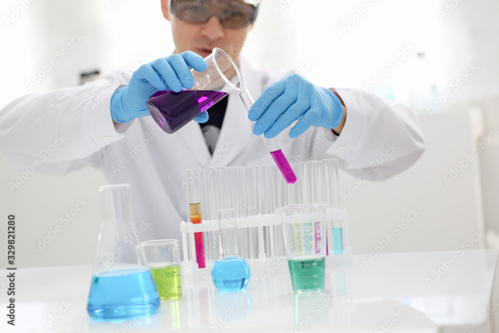 Male chemist holds test tube of glass in his hand overflows a liquid solution of potassium permanganate conducts an analysis reaction takes various versions of reagents using chemical manufacturing. Male chemist holds test tube of glass in his hand overflows a liquid solution of potassium permanganate conducts an analysis reaction takes various versions of reagents using chemical manufacturing.