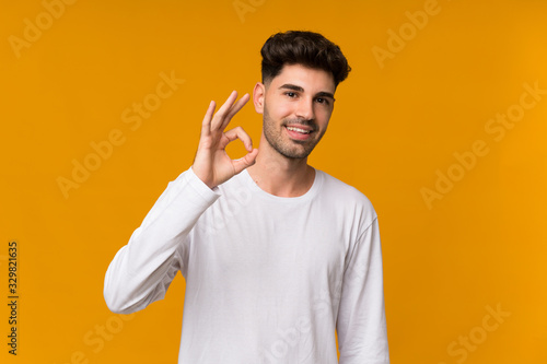 Young man over isolated orange background surprised and showing ok sign