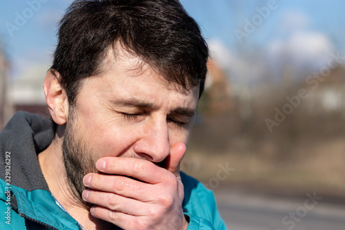 Coughing, sneezing or yawning squinting bearded man, covering his mouth with his hand on the street on a sunny day. Sick or tired man. Closeup view. Blurred background
