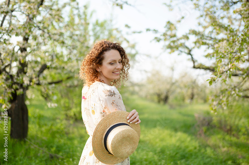 Young attractive woman with curly hair in a stylish wicker hat walks in a flowering green garden in spring day